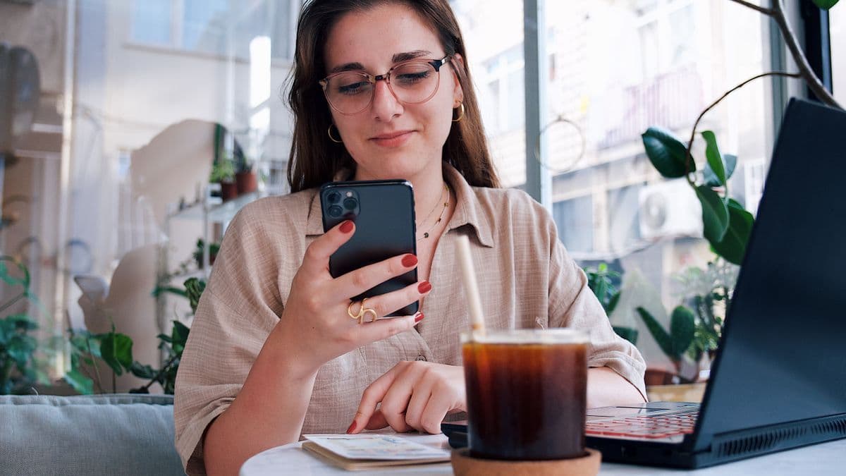 Telefonuna bakan, önünde dizüstü bilgisayar olan bir kadın
